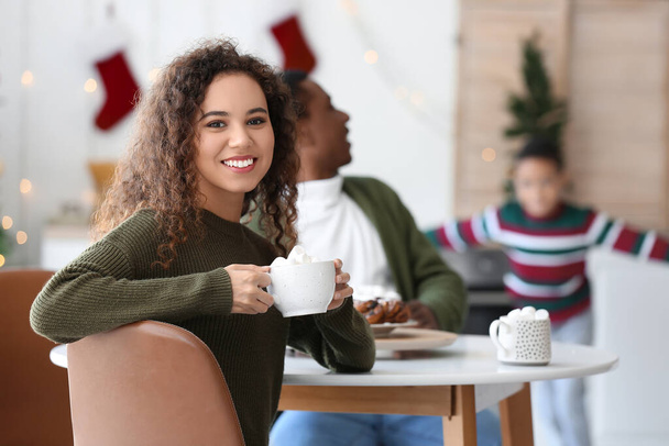 Young African-American woman with cup of hot chocolate and her family at home on Christmas eve - Фото, зображення