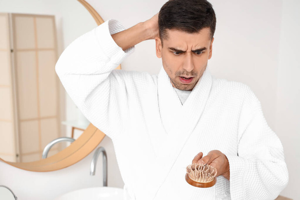 Hombre joven preocupado sosteniendo cepillo con el pelo caído en el baño - Foto, Imagen