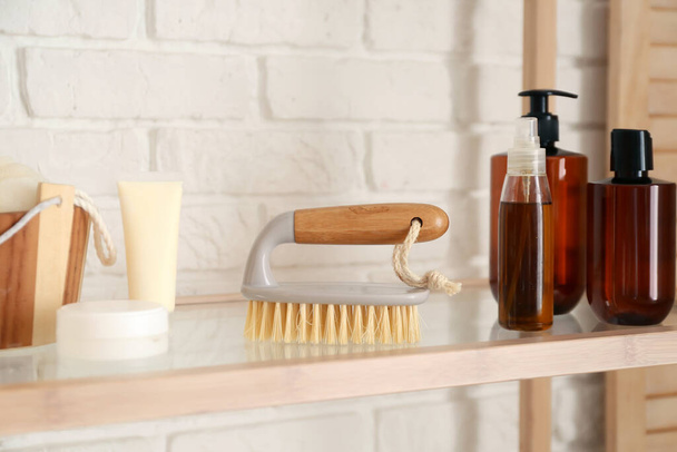Body massage brush and bottles with cosmetic products on shelf near white brick wall - Foto, Imagen