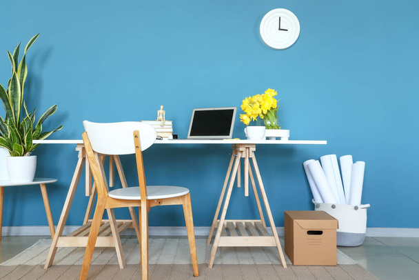 Lugar de trabajo con portátil moderno y jarrón con flores cerca de la pared azul en el interior de la habitación - Foto, imagen