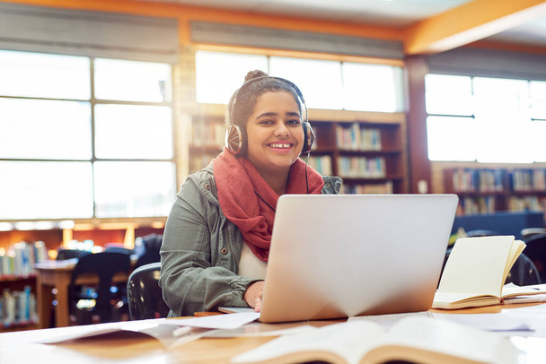 Η μουσική την αναγκάζει να παίζει. Πορτρέτο μιας χαρούμενης νεαρής φοιτήτριας που εργάζεται σε ένα φορητό υπολογιστή ακούγοντας μουσική με τα ακουστικά της μέσα σε μια βιβλιοθήκη. - Φωτογραφία, εικόνα