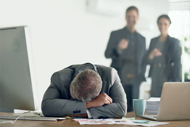 Pulling someone down wont help you reach the top. Shot of a dejected businessman lying on his arms at his desk while his colleagues mock him in the background. - Photo, image