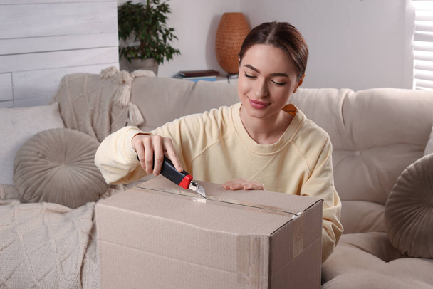 Young woman using utility knife to open parcel at home - Fotoğraf, Görsel