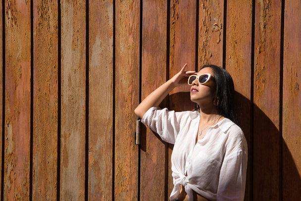 Молодая и красивая женщина, южноамериканка, в белой узловой рубашке и солнцезащитных очках, в профиль, на деревянной стене, на солнце. Красота, мода, отдых, путешествия. - Фото, изображение