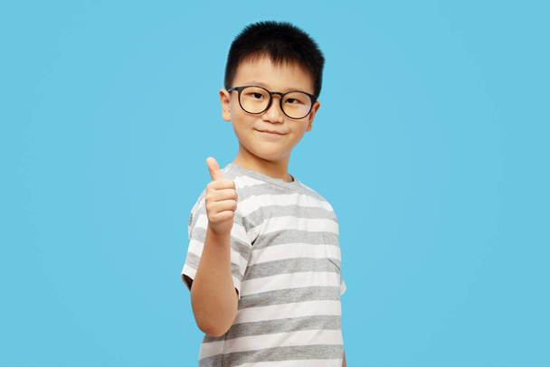 Chytrý kluk ukazuje náraz nahoru nosit brýle a tričko na modrém pozadí  - Fotografie, Obrázek