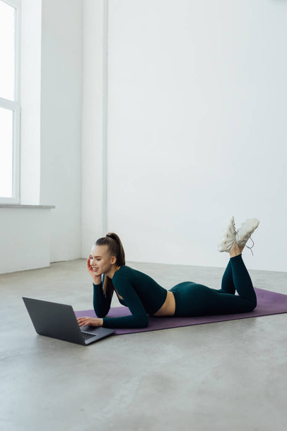 Ausbildungskonzept. Lächelnd fitte junge Frau in Sportkleidung, die sich mit Laptop am Fenster Fitnessvideos im Internet ansieht, im modernen Wohnzimmer mit minimalem Interieur oder Fitnessraum auf einer Yogamatte liegend - Foto, Bild