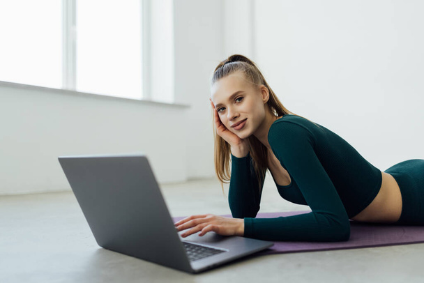 Training Concept. Smiling fit young woman in sports clothes watching fitness videos on internet using laptop near window, lying on floor yoga mat in the modern living room with minimal interior or gym - Photo, image
