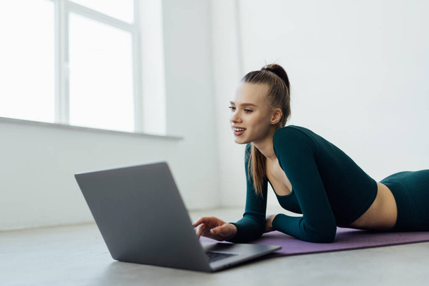 Képzési koncepció. Mosolygó fitt fiatal nő sportruhában néz fitness videók az interneten laptoppal ablak közelében, fekvő padló jóga szőnyeg a modern nappaliban, minimális belső vagy tornaterem - Fotó, kép