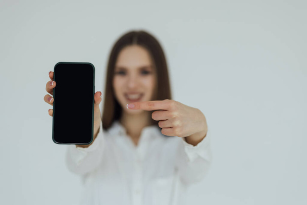 Happy young woman holding blank screen mobile phone and pointing finger over white background - Foto, Bild