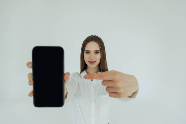 Retrato de una joven mujer de negocios emocionada señalando con el dedo el teléfono móvil en blanco aislado sobre fondo blanco
 - Foto, Imagen