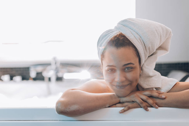 portrait of young attractive brazilian girl taking a bath in jacuzzi wearing bath towel on head - beautiful woman in spa treatments - smiling young woman relaxing in bathtub - Photo, Image