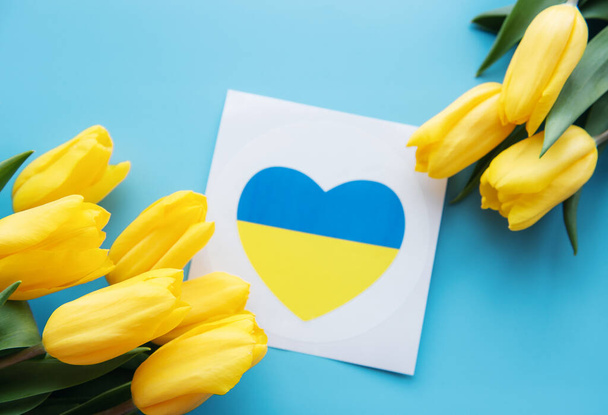 Card with a heart in the colors of the Ukrainian flag and yellow tulips on a blue background. Support for Ukraine - Фото, зображення