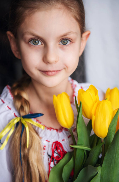 A little Ukrainian girl in traditional clothes holds a yellow tulips in her hands. Support for Ukraine. Stop the war. - Фото, изображение