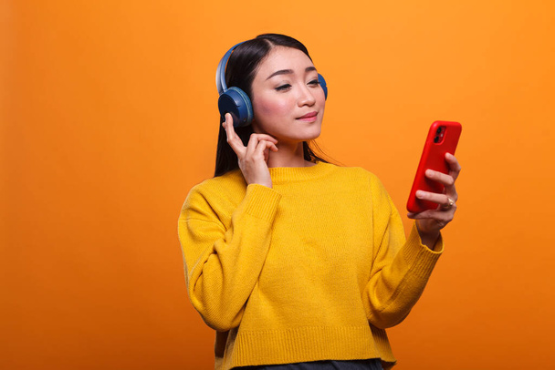 Relaxed friendly cute asian woman listening to music on audio streaming platform while using smartphone device. - Φωτογραφία, εικόνα