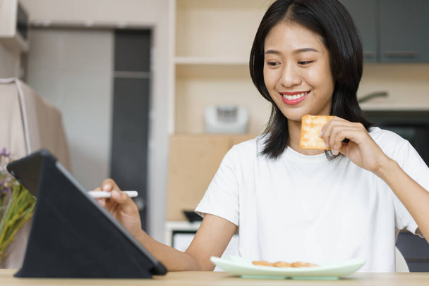Concepto de estilo de vida en casa, Mujer joven que usa tabletas y come galletas mientras está de ocio en casa. - Foto, Imagen