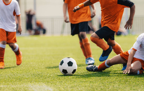 Grupo de niños multiétnicos jugando al fútbol. Chicos jóvenes corriendo tras la pelota de fútbol en el campo de fútbol de hierba. Niños en camisas de punto naranja y blanco. El jugador intenta hacer frente a una pelota de fútbol - Foto, Imagen