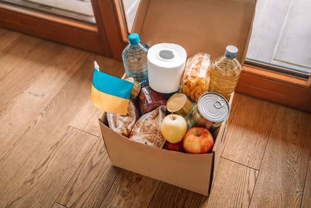  Paper full donation box of different grocery products for Ukraine children and people on wooden floor near the door. Donation Ukraine. Volunteering, Donations, charity refugees war. - Foto, Bild