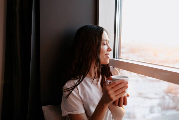 Close up retrato de menina encantadora feliz com cabelo escuro está segurando café e olhando para a janela com sorriso feliz. Novo dia em casa com sol. Sozinho em casa. Foto de alta qualidade - Foto, Imagem