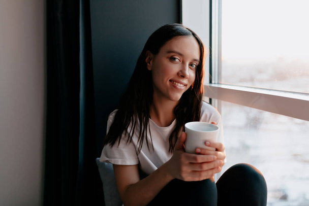 appealing girl with long wavy hair and wonderful smile wearing white t-shirt is sitting near the window with cup of coffee in the morning . High quality photo - Photo, Image