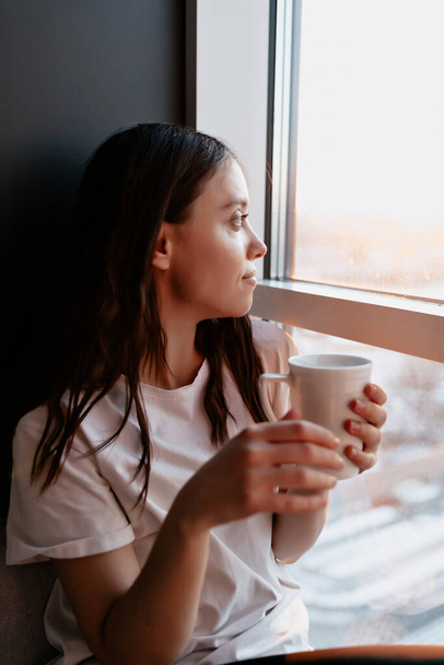 Close up portrait of dark-haired girl in white t-shirt is holding cup with coffee and looking in the window with charming smile in the morning . High quality photo - 写真・画像