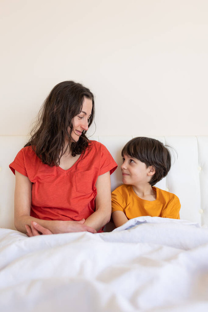 Un enfant avec maman s'assoit sur le lit, une femme et un garçon en pyjama, une femme étreint son fils, matin avec maman - Photo, image