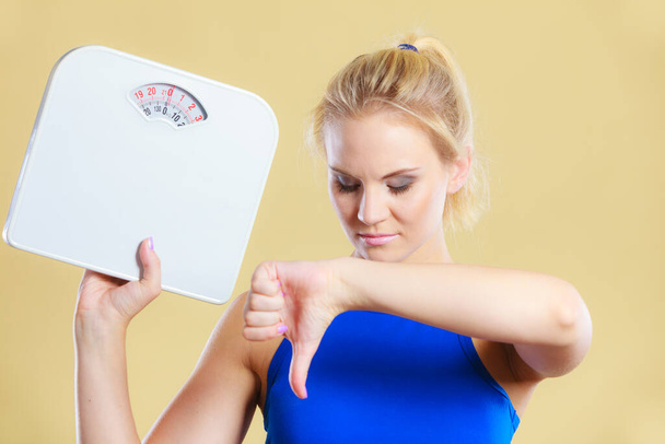 Frustrated sad blonde girl holding scales, making thumb down gesture sign. Weight gain, time for slimming weightloss, diet. - Foto, afbeelding