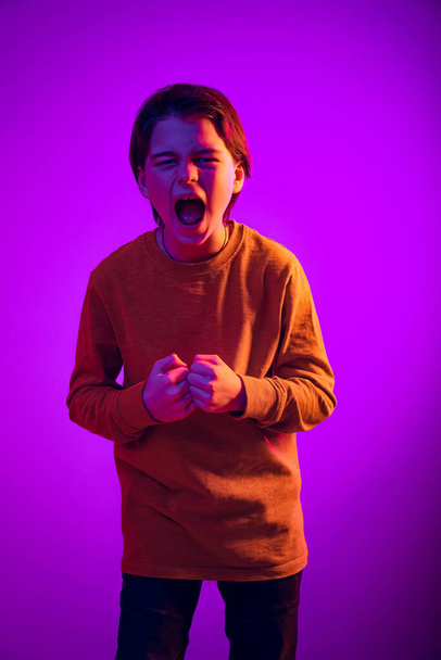 Portrait of emotive boy, child shouting, screaming isolated over purple background in neon light - Photo, image