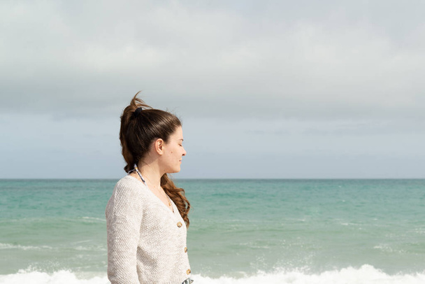 Vue du profil d'une jeune femme aux yeux fermés profitant d'un rayon de soleil lors d'une journée orageuse avec la mer en arrière-plan. Concept inspirant. - Photo, image