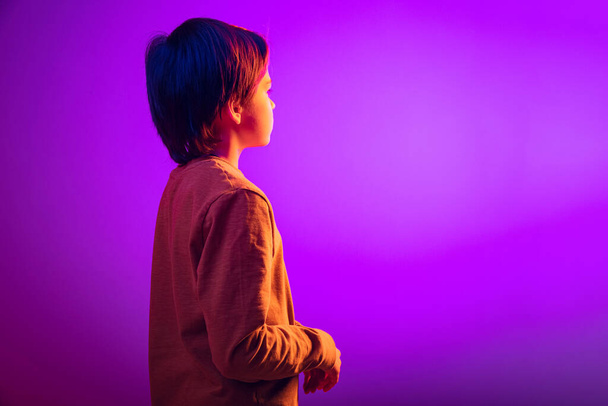 Side view portrait of boy, child dreamily looking away isolated over purple background in neon light - Фото, зображення