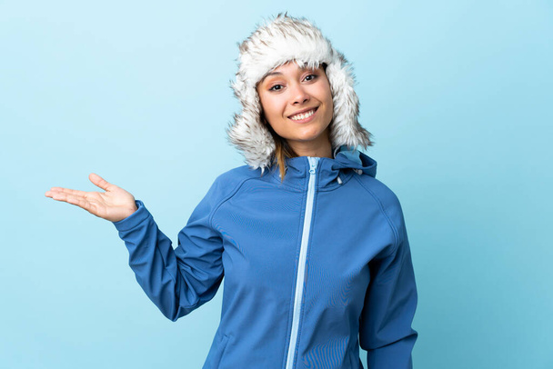 Молодая уругвайская девушка в зимней шляпе изолирована на синем фоне, держа копирайт на ладони. - Фото, изображение