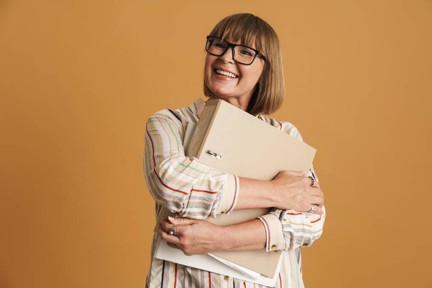 Senior woman wearing eyeglasses smiling and posing with folders isolated over beige wall - Φωτογραφία, εικόνα