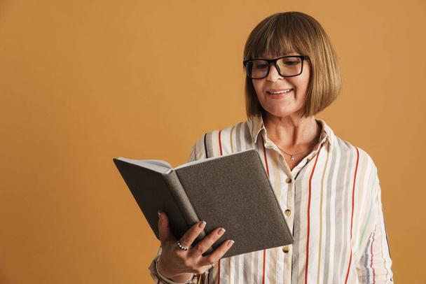 Senior woman wearing eyeglasses smiling while posing with planner isolated over beige wall - Φωτογραφία, εικόνα
