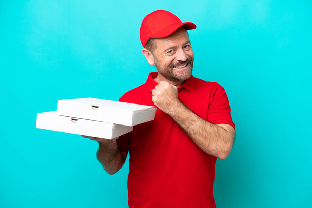 Repartidor de pizza con uniforme de trabajo recogiendo cajas de pizza aisladas sobre fondo azul celebrando una victoria - Foto, Imagen