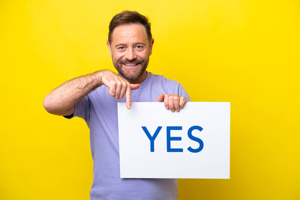 Middle age caucasian man isolated on yellow background holding a placard with text YES and pointing it - 写真・画像