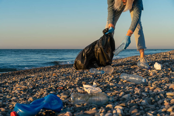 Młoda kobieta zbiera plastikowe, gumowe śmieci w czarnym worku na śmieci na żwirowej plaży nad morzem o zachodzie słońca. Puste zużyte plastikowe butelki. Zanieczyszczenie środowiska na wybrzeżu Morza Czarnego - Zdjęcie, obraz