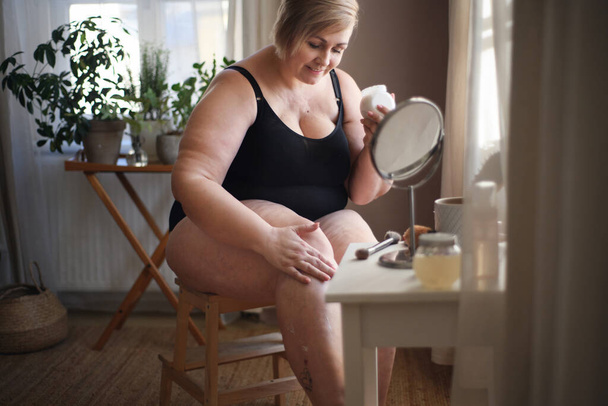Ylipainoinen nainen istuu ja levittää kermaa jalkoihinsa kotona, itsehoidon käsite. - Valokuva, kuva