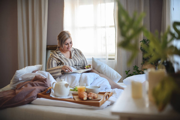 Happy overweight woman with book having breakfast in bed at home. - Foto, afbeelding