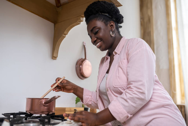 Elégedett afrikai nő rózsaszín ruhában főzés a konyhában - Fotó, kép