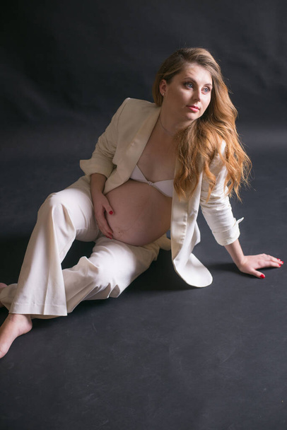 Young cute pregnant woman with long red hair in a white trouser suit on a black background. Happy motherhood. Mothers Day - Фото, зображення