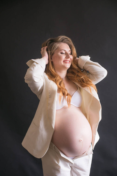Young cute pregnant woman with long red hair in a white trouser suit on a black background. Happy motherhood. Mothers Day - Foto, afbeelding