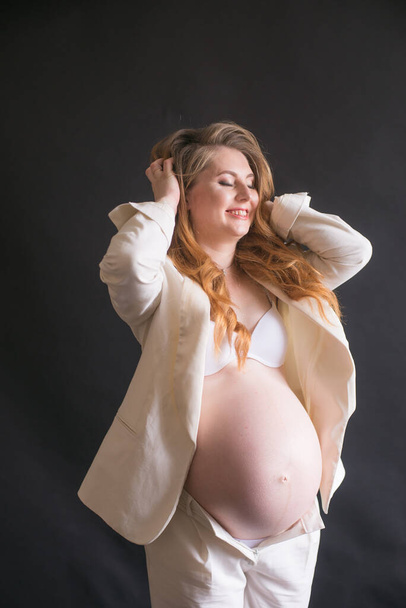 Young cute pregnant woman with long red hair in a white trouser suit on a black background. Happy motherhood. Mothers Day - Photo, Image