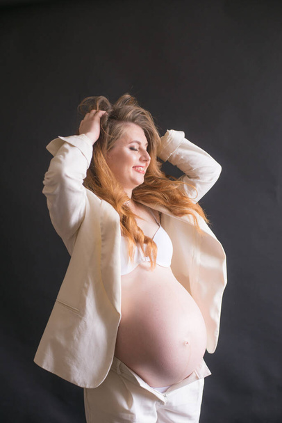 Young cute pregnant woman with long red hair in a white trouser suit on a black background. Happy motherhood. Mothers Day - Foto, Imagem