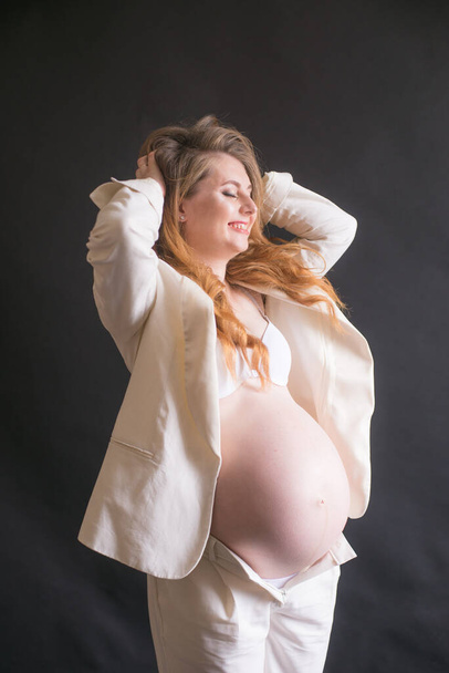 Young cute pregnant woman with long red hair in a white trouser suit on a black background. Happy motherhood. Mothers Day - Fotografie, Obrázek
