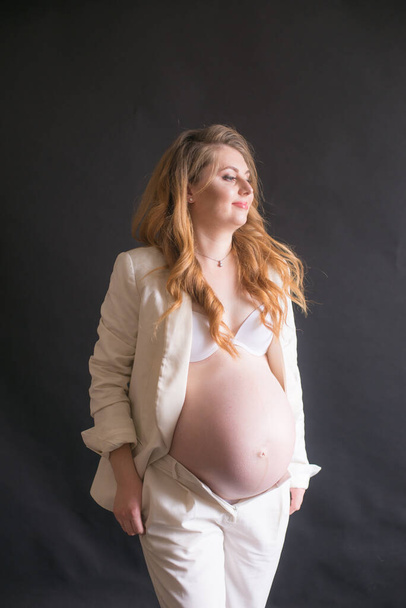 Young cute pregnant woman with long red hair in a white trouser suit on a black background. Happy motherhood. Mothers Day - Photo, Image