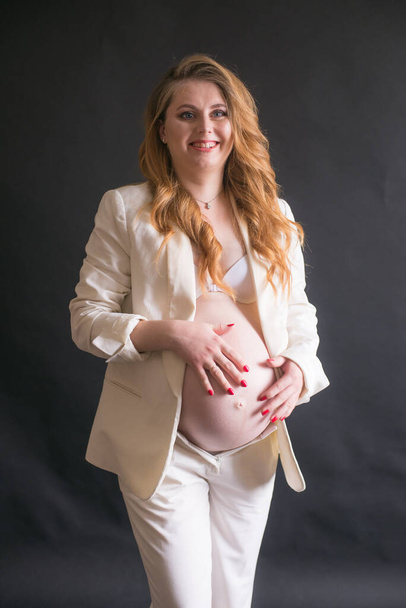 Young cute pregnant woman with long red hair in a white trouser suit on a black background. Happy motherhood. Mothers Day - Fotó, kép