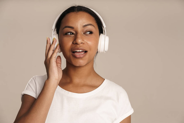 Молода чорна жінка співає під час прослуховування музики з навушниками ізольовано над білою стіною
 - Фото, зображення