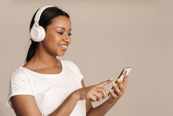 Jovem mulher negra sorrindo enquanto usa celular e fones de ouvido isolados sobre a parede branca - Foto, Imagem