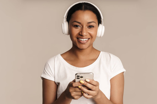 Joven mujer negra sonriendo mientras usa el teléfono celular y los auriculares aislados sobre la pared blanca - Foto, imagen
