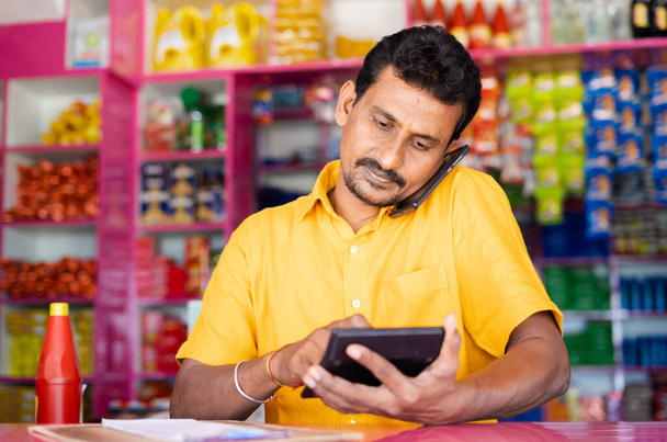 Kirana or grocery marchant calculating order price while taking on mobile phone from customer - concept of successful small business, communication and retail store. - Photo, image
