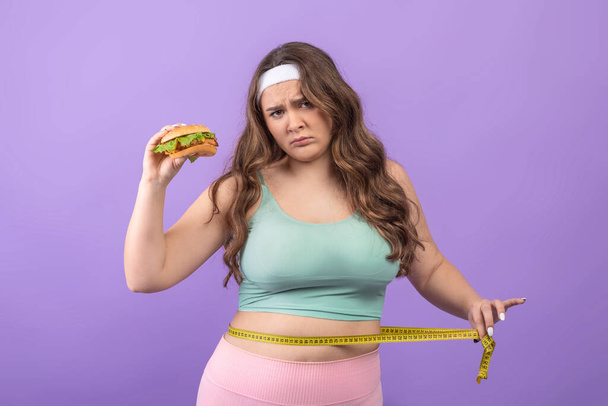 Triste europeo joven hambriento más tamaño femenino en ropa deportiva con hamburguesa en la mano, mide su cintura con cinta métrica - Foto, Imagen
